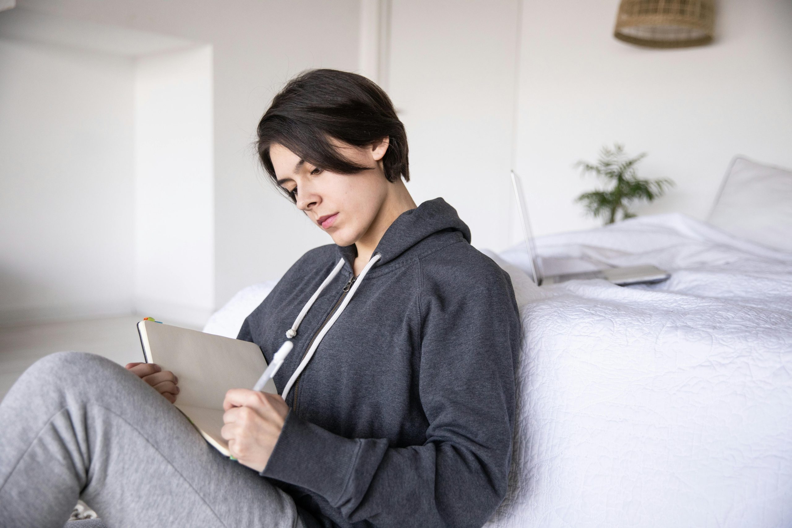 A young woman sits by a bed, writing in a notebook in a cozy bedroom.